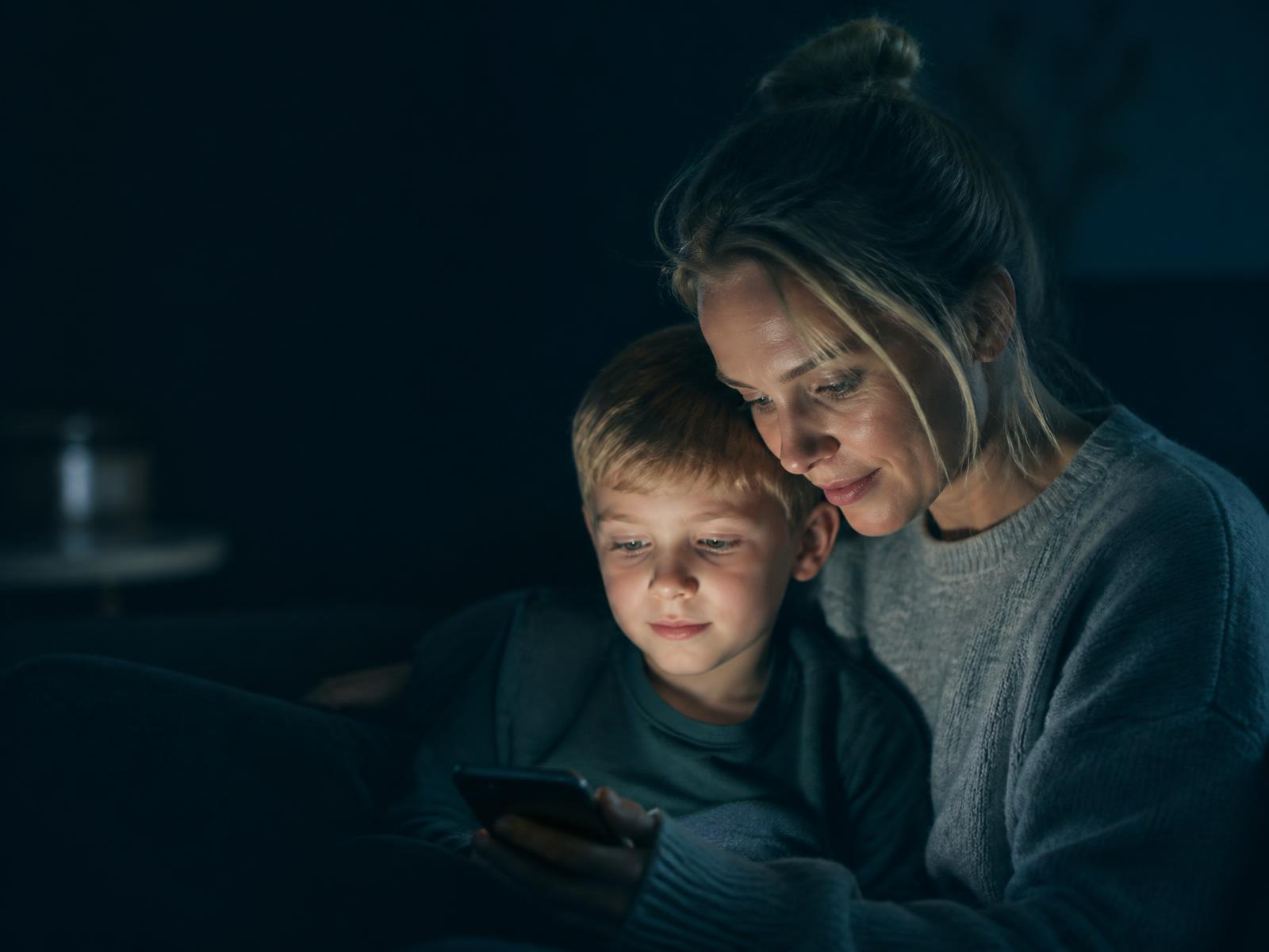 Förälder och barn tittar tillsammans på en telefon i ett mörkt vardagsrum